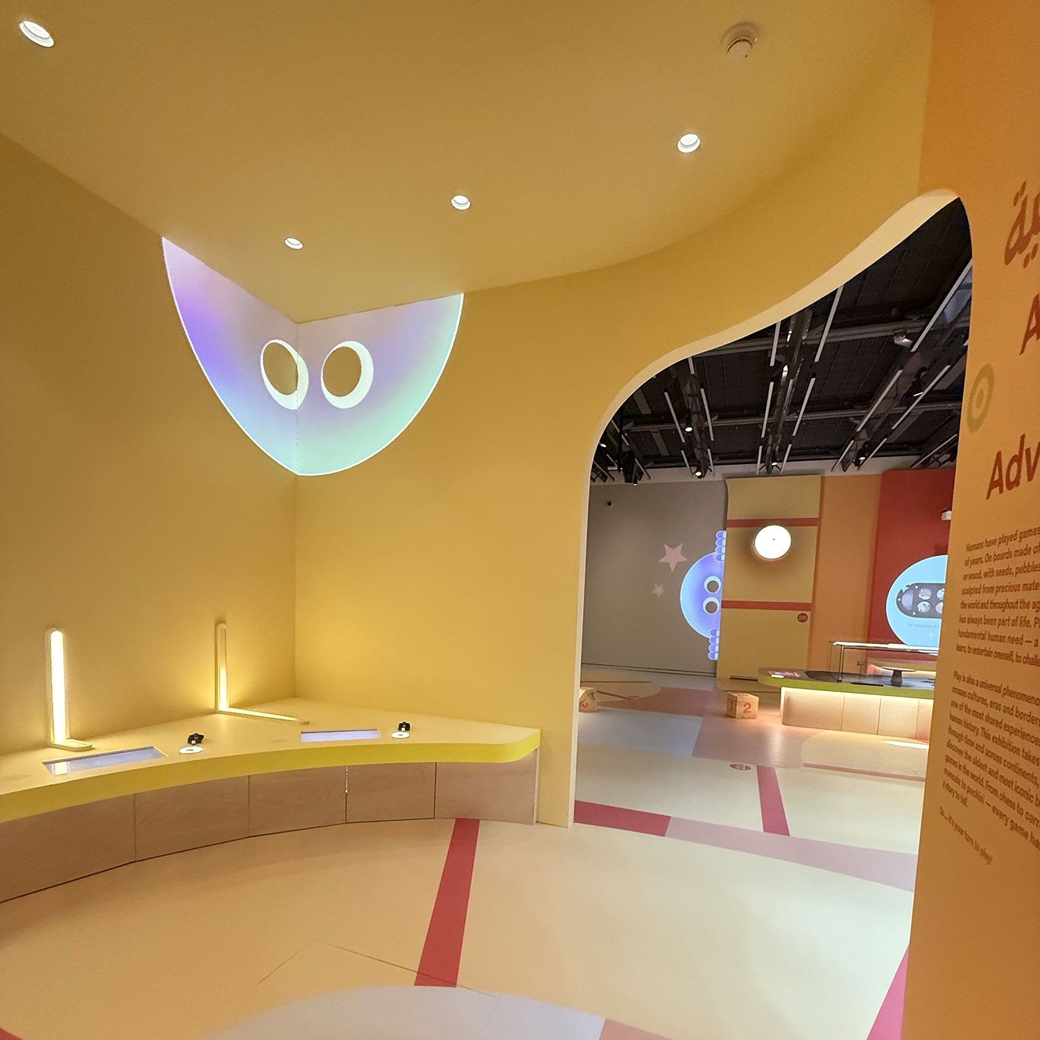 L'image montre un espace intérieur aux murs de teintes jaunes, avec un éclairage doux et des éléments de design moderne. On y voit des courbes et des formes géométriques sur le sol, qui semblent être un mélange de couleurs pastel. Des affichages lumineux projettent des formes ou des visages stylisés sur les murs. On note aussi des zones de travail ou d'exploration, avec des tables et des sièges. L'ensemble crée une atmosphère accueillante et ludique.