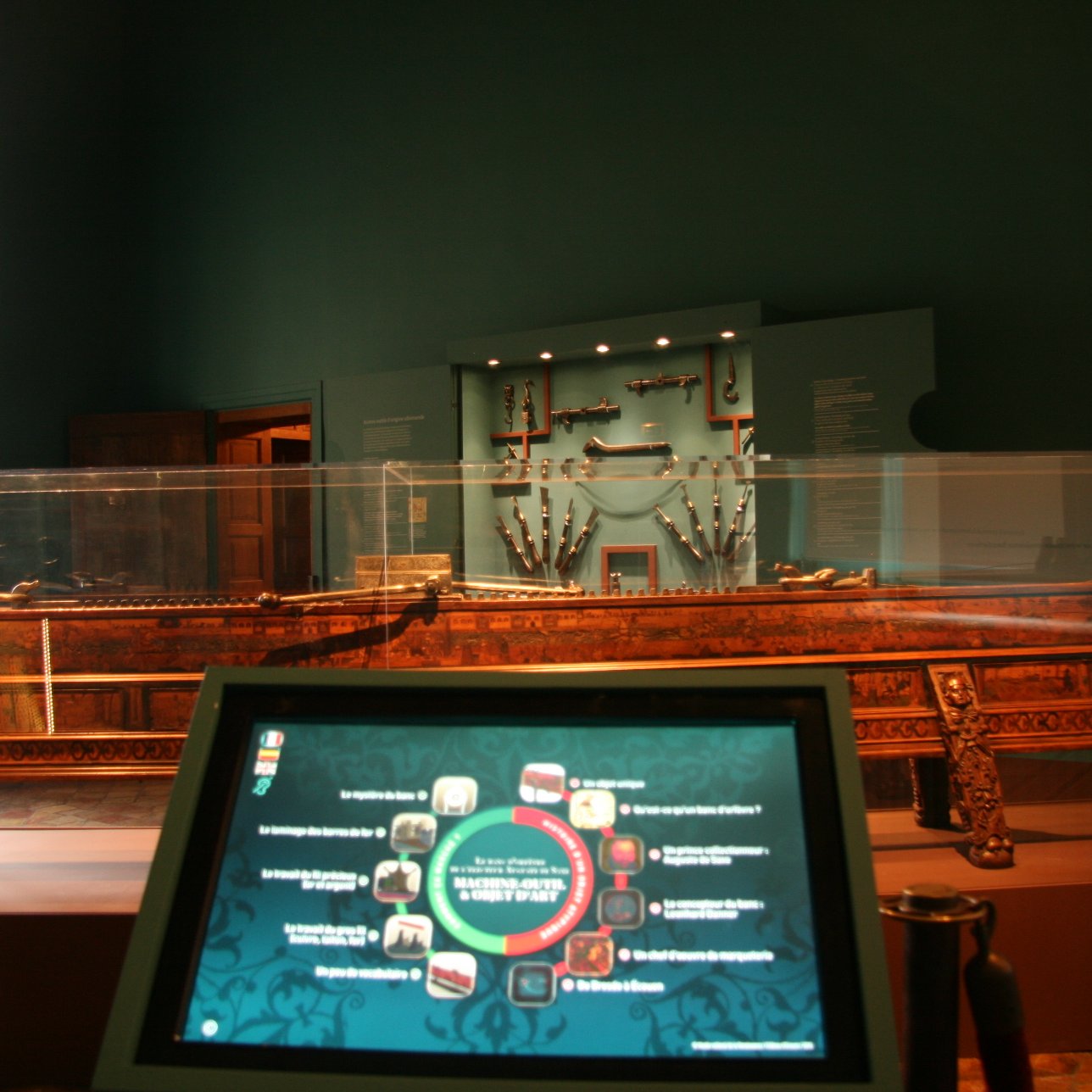 L'image montre un intérieur de musée où l'on peut voir un grand meuble en bois décoré, probablement une pièce d'artisanat ou un objet historique exposé dans une vitrine transparente. À l'arrière-plan, un mur est décoré avec plusieurs objets accrochés, qui pourraient être des armes ou des outils anciens. Au premier plan, il y a un écran interactif qui semble fournir des informations sur l'exposition. L'ambiance est calme, avec des murs peints dans une teinte sombre qui met en valeur les objets exposés.