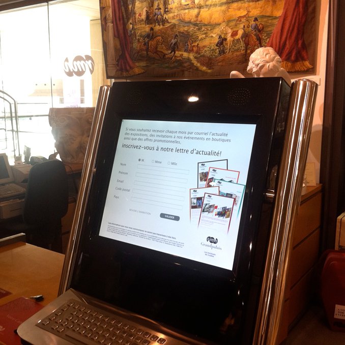 L'image montre un kiosque informatique moderne placé sur un comptoir. L'écran tactile est affiché, avec un formulaire pour s'inscrire à une lettre d'actualité, indiquant où entrer des informations. On aperçoit également des objets en arrière-plan, tels que des vitrines et une tapisserie. La conception du kiosque semble élégante et contemporaine.
