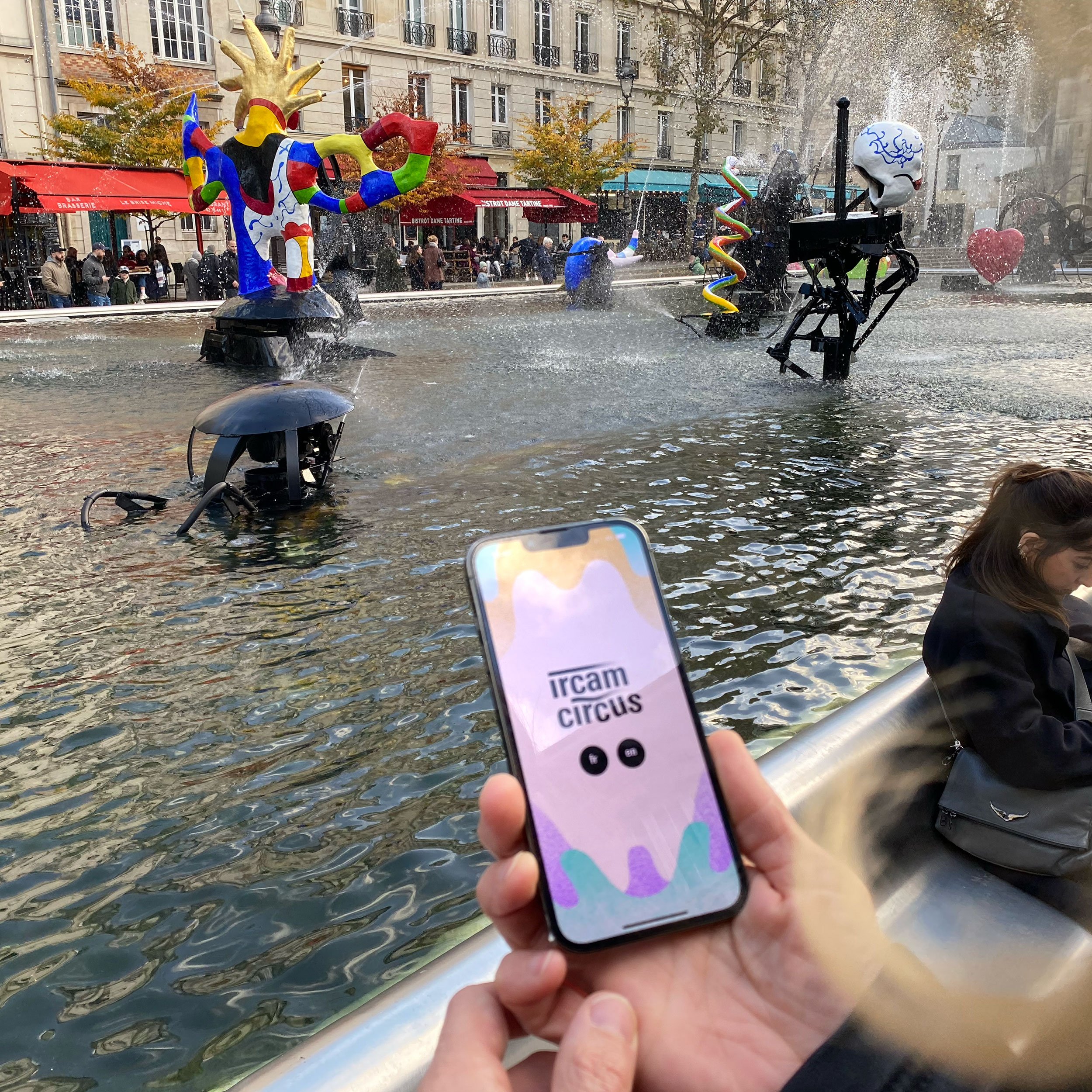 L'image montre une scène animée d'une place publique avec une fontaine au centre. On peut voir plusieurs sculptures colorées disposées autour de la fontaine. À l'avant-plan, une personne tient un smartphone affichant l'application "ircam circus". À droite, une autre personne est assise, probablement en train d'observer la scène. L'ambiance semble vivante et artistique, avec des couleurs vives et des éléments d'eau en mouvement.