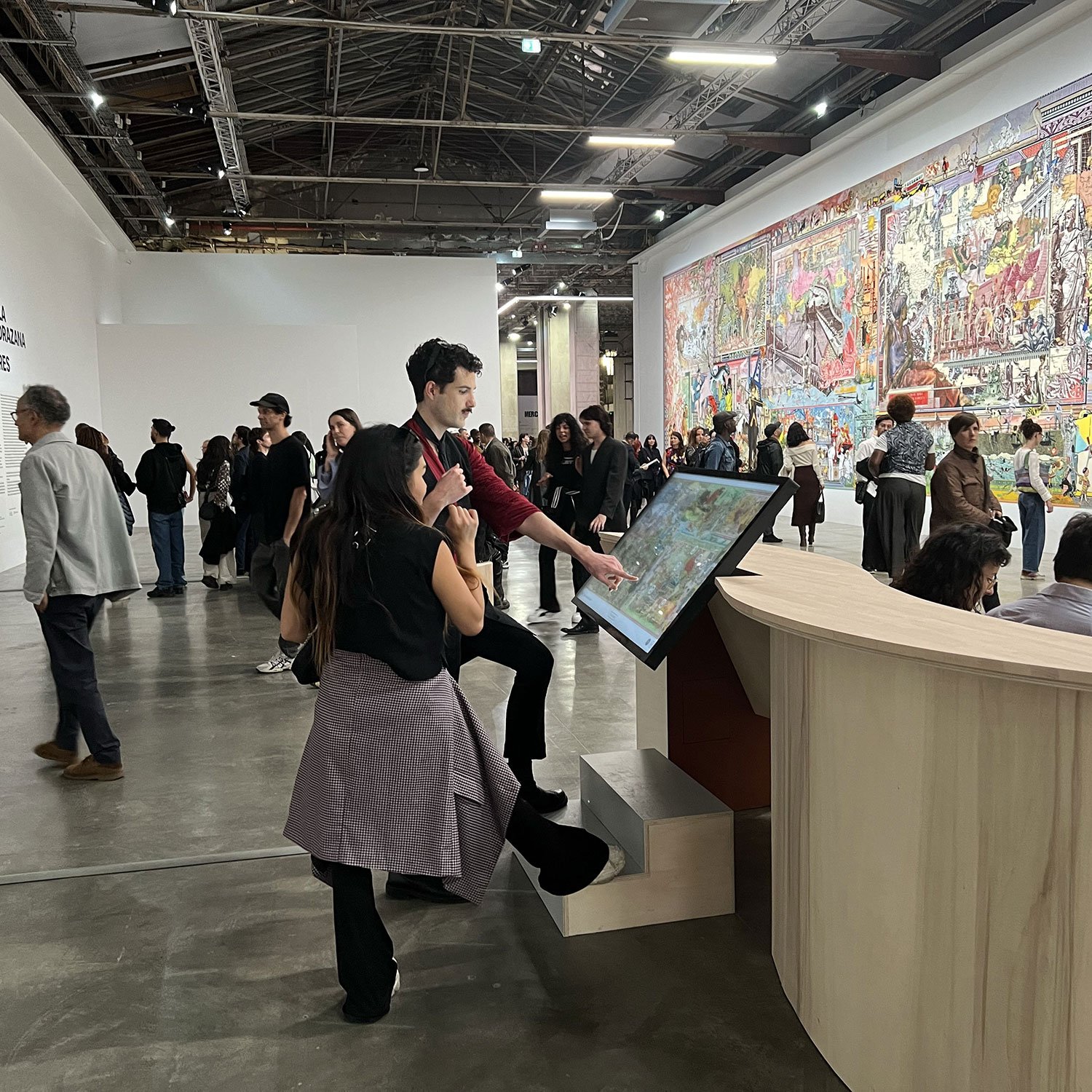 L'image montre une exposition artistique dans un grand espace. On peut voir un mur rempli de panneaux illustrés, avec une œuvre colorée qui attire l'attention. Au milieu, un groupe de personnes interagit avec un écran tactile, tandis que d'autres spectateurs explorent les environs. L'ambiance est animée, avec des visiteurs de tous âges qui découvrent les œuvres exposées. La lumière est vive et met en valeur les détails du décor.