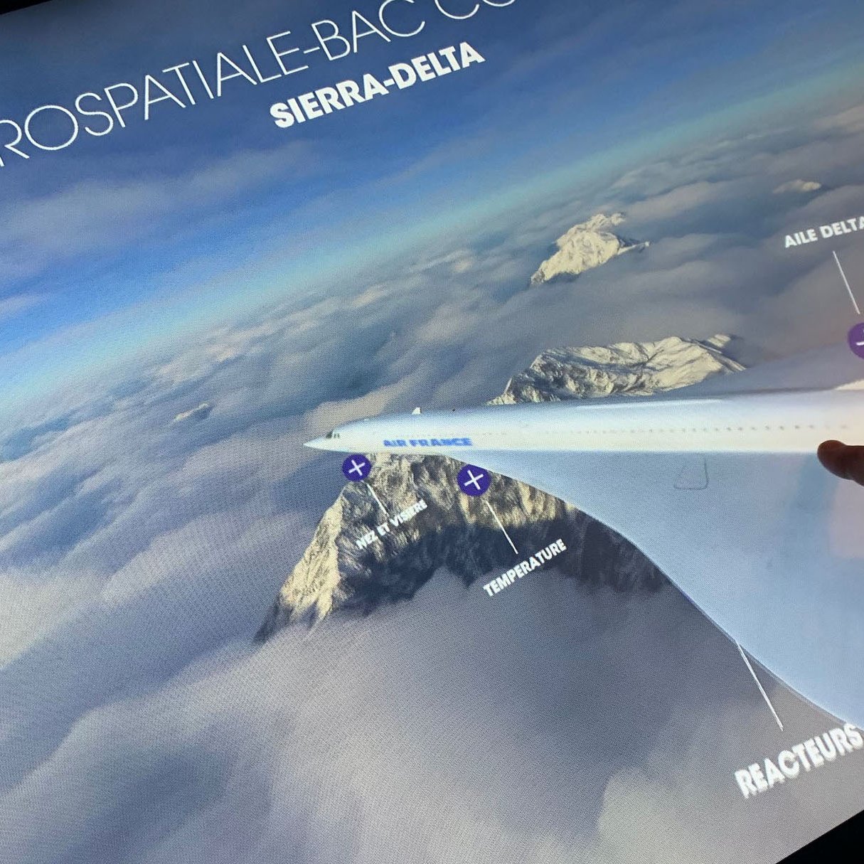 L'image montre un écran interactif qui présente un modèle d'avion dans un environnement aérien. On peut voir des montagnes sous des nuages, et un doigt pointe vers l'aile de l'appareil, où des annotations étiquettent des éléments comme "Aile Delta", "Réacteur" et "Température". Le titre indique qu'il s'agit d'une présentation sur l'aéronautique, plus précisément sur le modèle "Sierra-Delta".