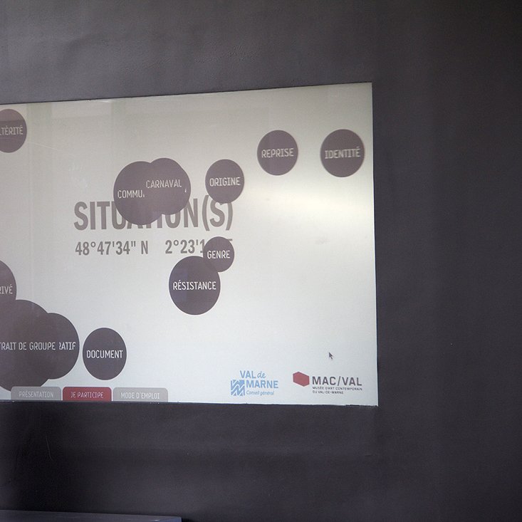 L'image montre un écran affichant le mot "SITUATION(S)" accompagné de plusieurs mots-clés en noir, tels que "IDENTITÉ," "DOCUMENT," et "RÉSISTANCE." Ces mots sont disposés de manière désordonnée, créant une sorte de carte mentale. En arrière-plan, on peut voir une personne dont seule la silhouette est visible, suggérant qu'elle regarde l'écran. L'environnement semble moderne et épuré.