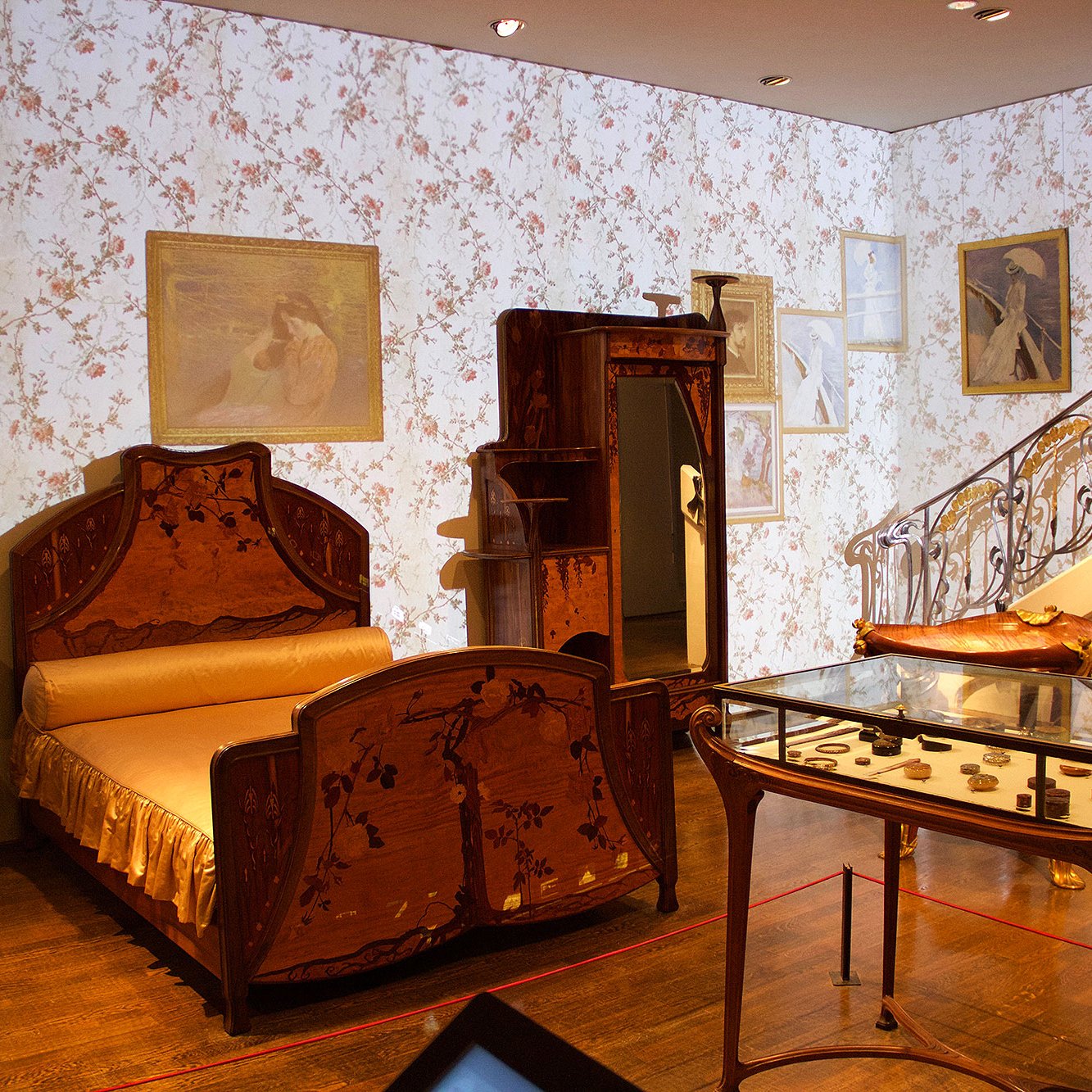 L'image montre une chambre d'époque avec un style décoratif élégant. On y voit un lit en bois orné de motifs floraux, une armoire au design similaire, et une table en verre reposant sur un sol en bois. Les murs sont recouverts d'un papier peint à fleurs, et des tableaux sont accrochés, contribuant à l'ambiance artistique de la pièce. Un escalier avec une balustrade ornée se trouve à l'arrière-plan, ajoutant une touche architecturale à l'ensemble. La lumière douce et les éléments décoratifs évoquent une atmosphère nostalgique et raffinée.