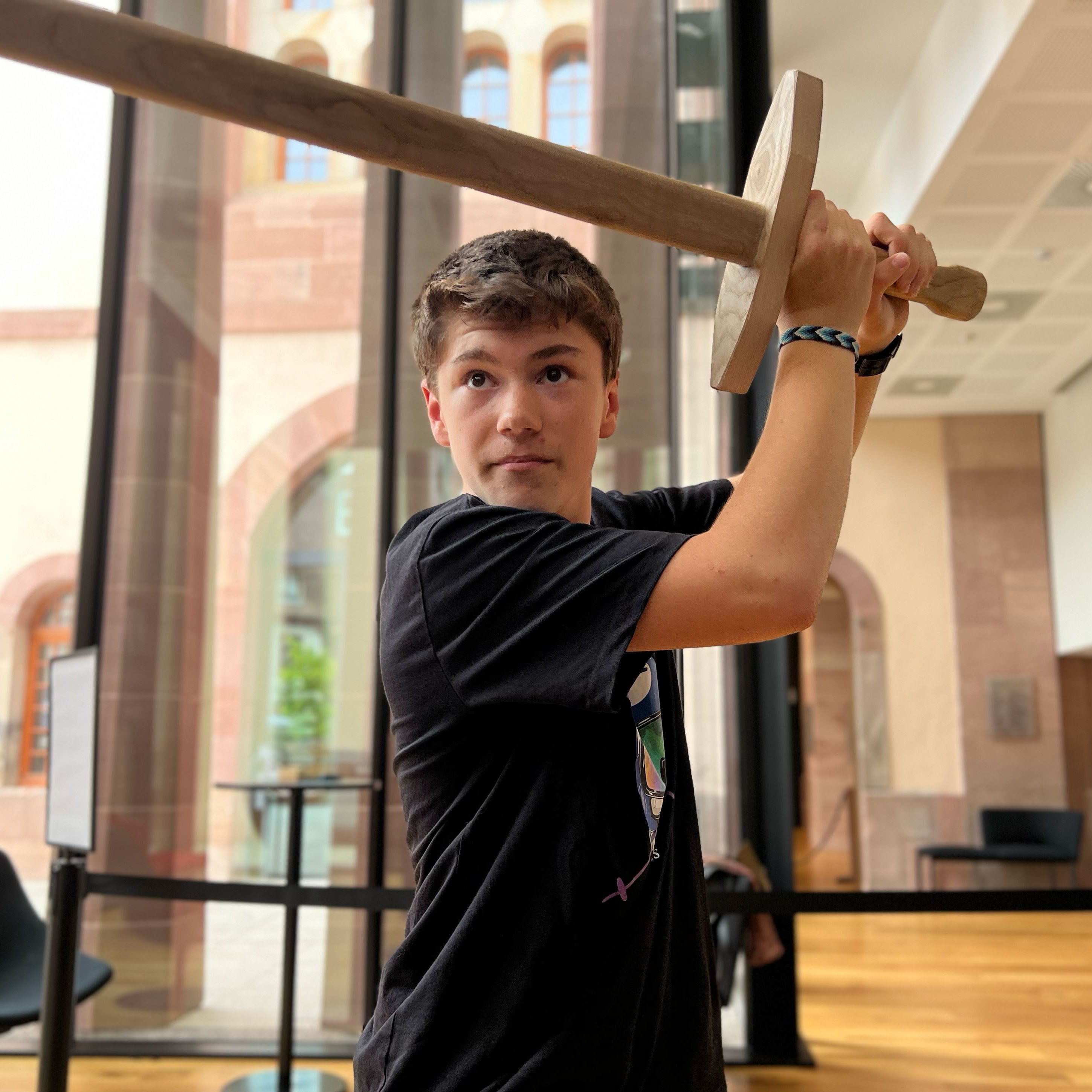 L'image montre un jeune homme levant une épée en bois. Il se tient dans un espace intérieur, probablement dans un musée ou un bâtiment historique, avec de grandes fenêtres et un design moderne. Le jeune homme porte un t-shirt noir et a une expression concentrée, semblant prêt à se battre ou à s'entraîner. L'ambiance générale est dynamique et engageante.