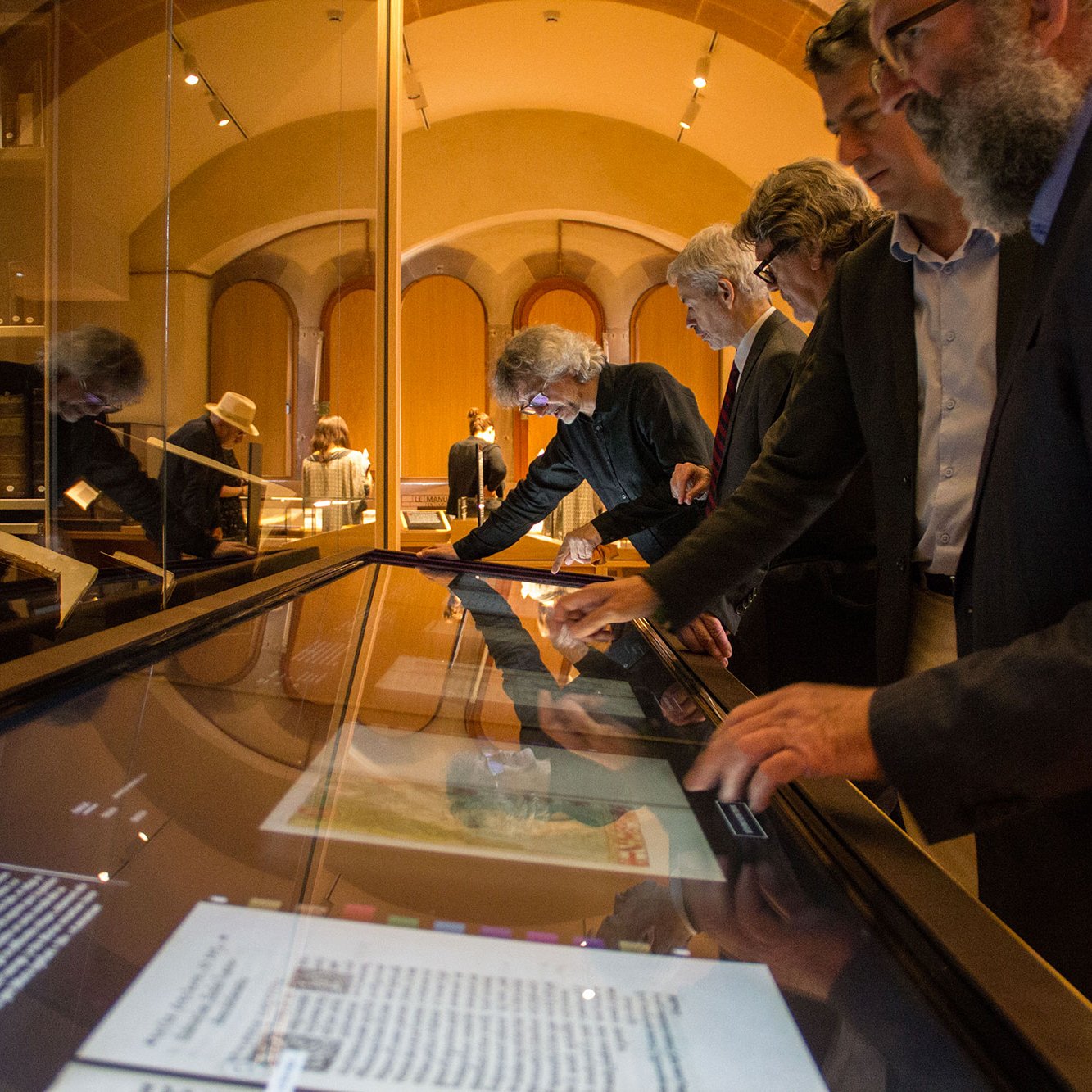 L'image montre un groupe de personnes observant attentivement des expositions dans un musée. Elles sont penchées sur un écran interactif ou une table d'exposition qui affiche des documents ou des images. L'ambiance semble studieuse, et la lumière douce met en valeur l'architecture intérieure du musée. Les visiteurs semblent engagés dans leur exploration, échangeant des idées et des remarques.