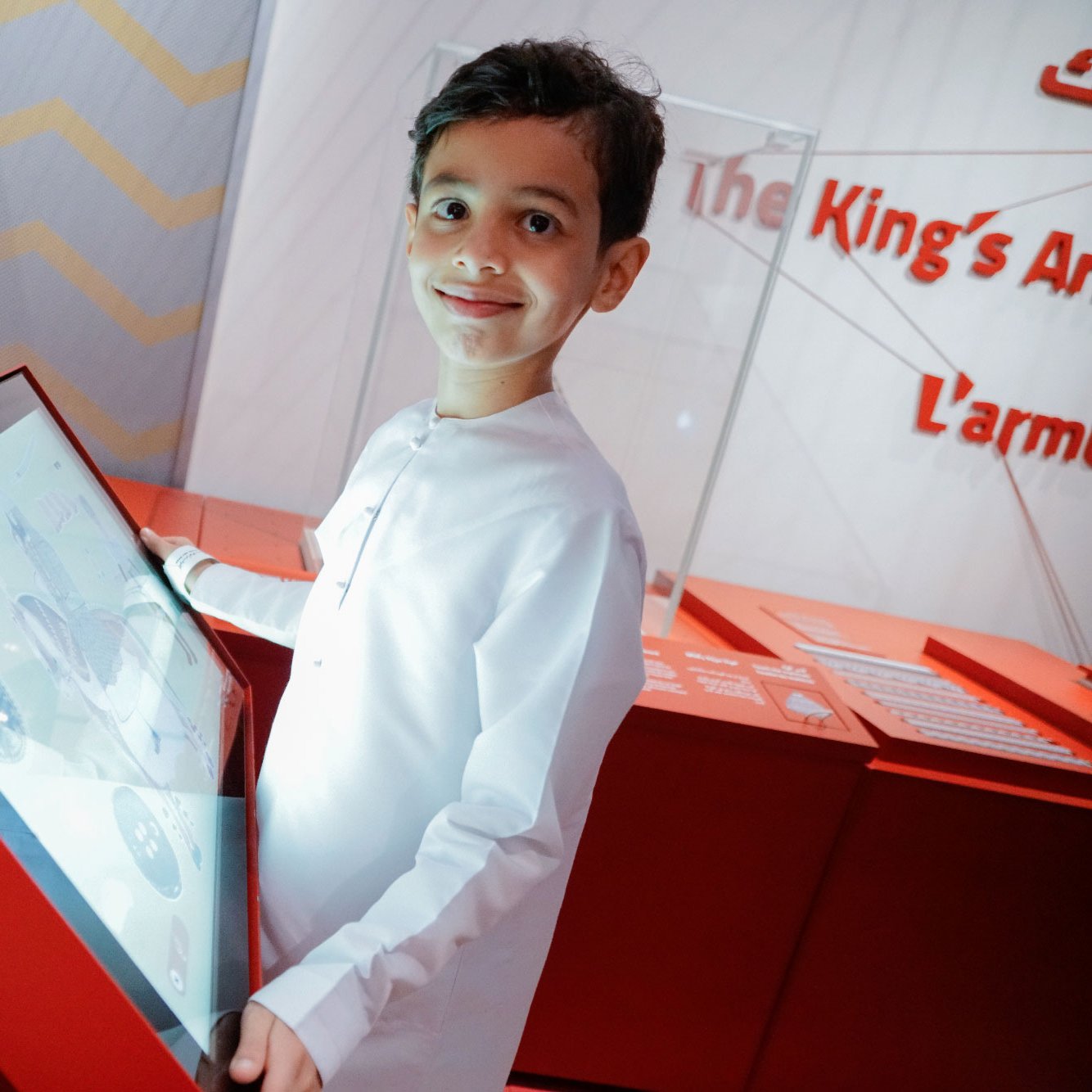 L'image montre un jeune garçon souriant, vêtu d'une tenue traditionnelle blanche. Il se tient près d'un écran tactile rouge, sur lequel il semble interagir avec du contenu. En arrière-plan, on peut voir des éléments de décoration moderne, créant une atmosphère accueillante et éducative. L'ensemble de la scène dégage une ambiance d'apprentissage et de découverte.