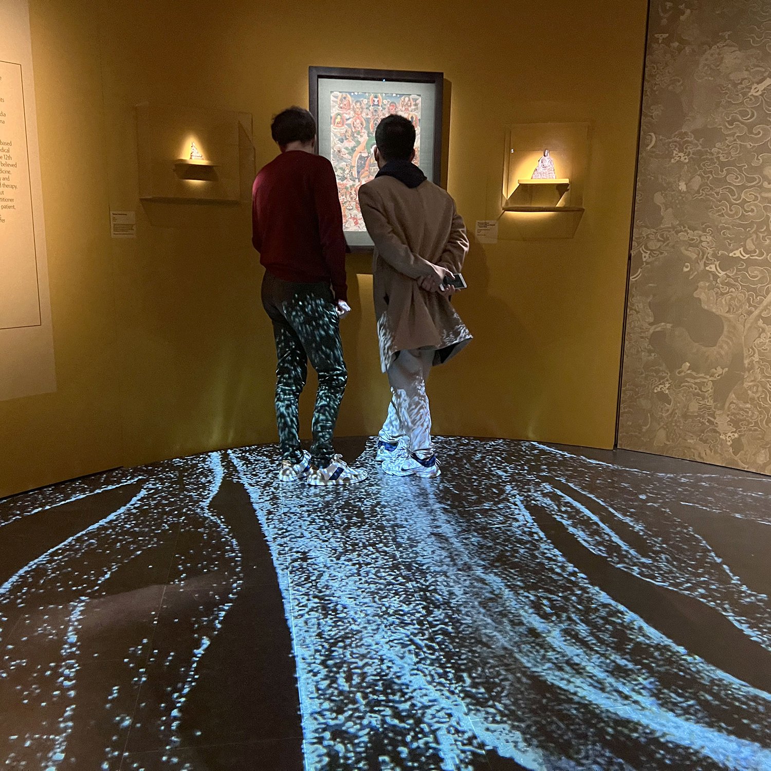 L'image montre deux personnes se tenant debout dans un espace d'exposition. Ils regardent attentivement une œuvre d'art affichée sur le mur. Le sol est recouvert d'une projection qui crée un effet visuel évoquant de l'eau ou des vagues. Les murs sont de couleur jaune, et il y a des éclairages discret sur les côtés qui mettent en valeur l'exposition. L'ambiance est à la fois contemplative et immersive.