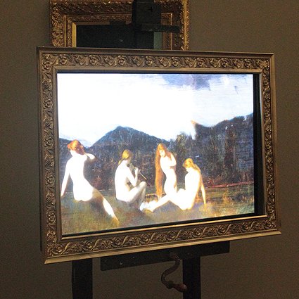 L'image semble représenter une œuvre d'art en format numérique, encadrée dans un cadre orné. On peut y voir des figures humaines, probablement nues, disposées dans un paysage avec des montagnes en arrière-plan. Les personnages sont assis ou posant de manière contemplative, suggérant une atmosphère paisible. La qualité de l'image peut donner l'impression d'un tableau classique, mais avec une touche moderne grâce à l'écran numérique.