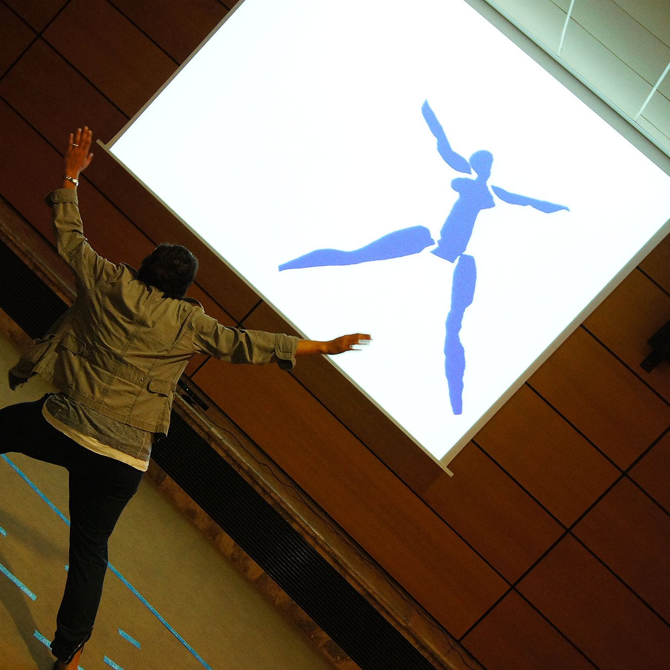 L'image montre une personne en mouvement, peut-être en train de danser ou de faire une performance, se tenant devant un grand écran. Sur cet écran, on voit une silhouette bleue représentant une figure humaine stylisée. L'environnement semble être une salle de réunion ou un espace de présentation, avec des murs en bois et un sol clair. La personne semble interagir avec l'image projetée, ajoutant une dynamique visuelle à la scène.