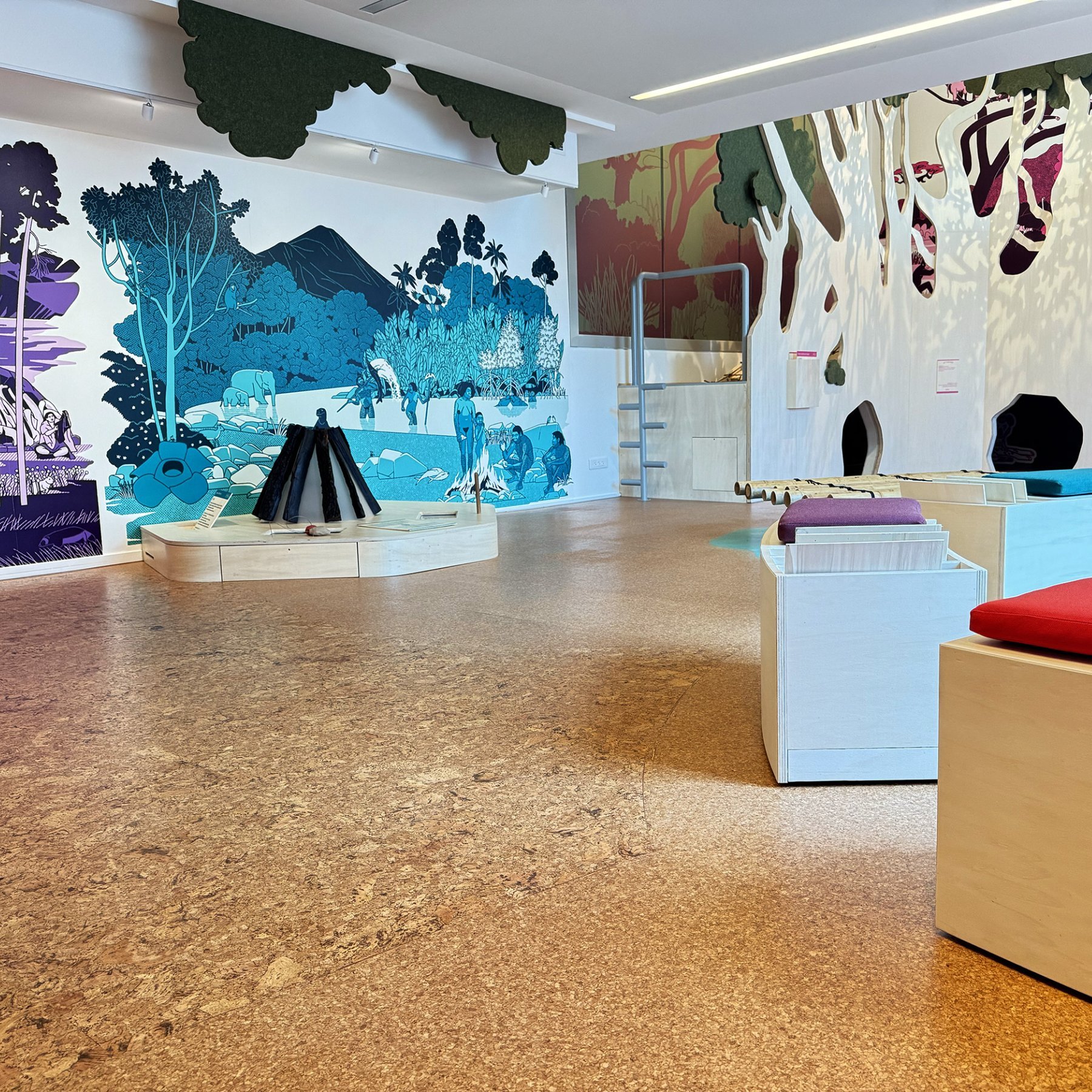 L'image montre un espace intérieur qui semble être une salle d'exposition ou un espace éducatif. Les murs sont décorés de grandes fresques colorées représentant des paysages et des scènes de nature, avec des arbres, des collines et des éléments animés. Au centre de la pièce, il y a plusieurs bancs en bois de différentes formes et couleurs, et une installation qui rappelle un feu de camp, posé au sol. L'éclairage est ample et met en valeur l'ambiance conviviale et accueillante de cet espace.