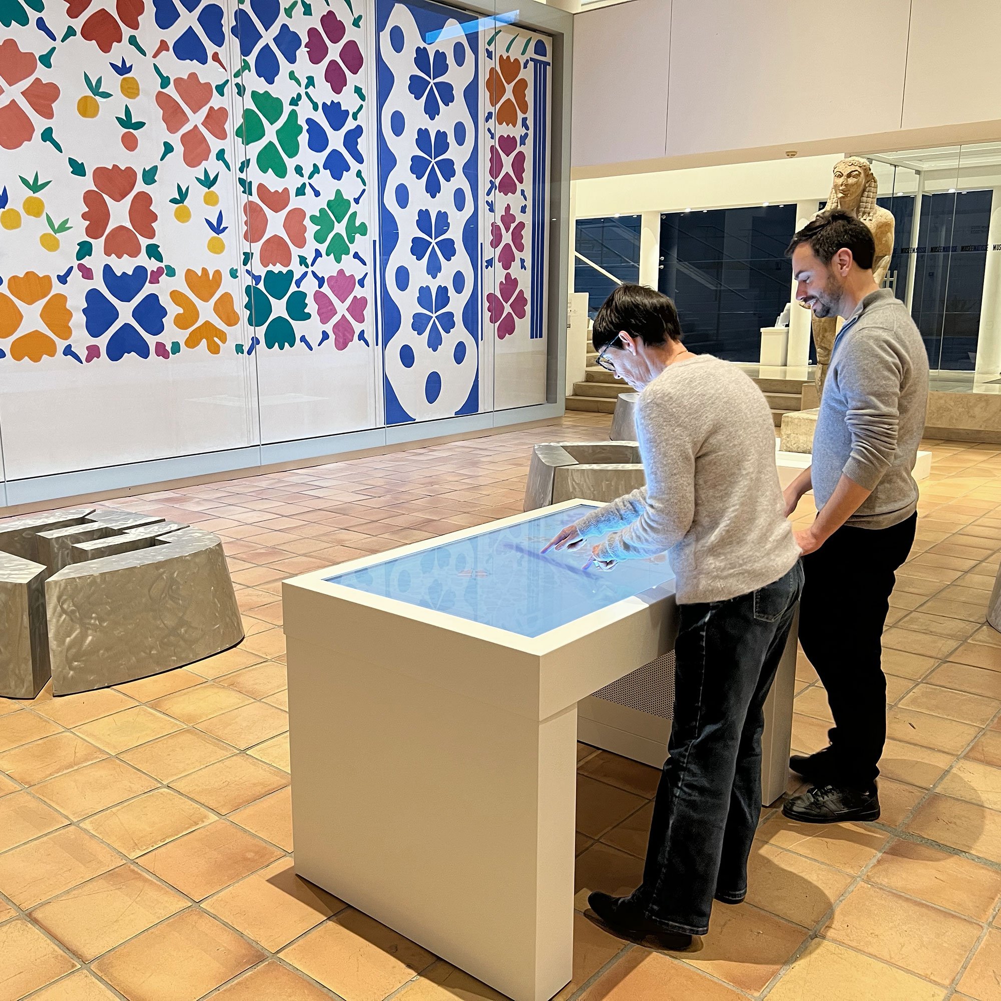 L'image montre une salle d'exposition d'un musée. Deux personnes se tiennent devant un écran interactif en touchant son interface. En arrière-plan, un grand mur est décoré de motifs colorés en forme de fleurs et de feuilles. Il y a également des sculptures ou des objets artistiques disposés dans la salle. Le sol est carrelé avec des carreaux en terre cuite, ajoutant une atmosphère chaleureuse à l'environnement.
