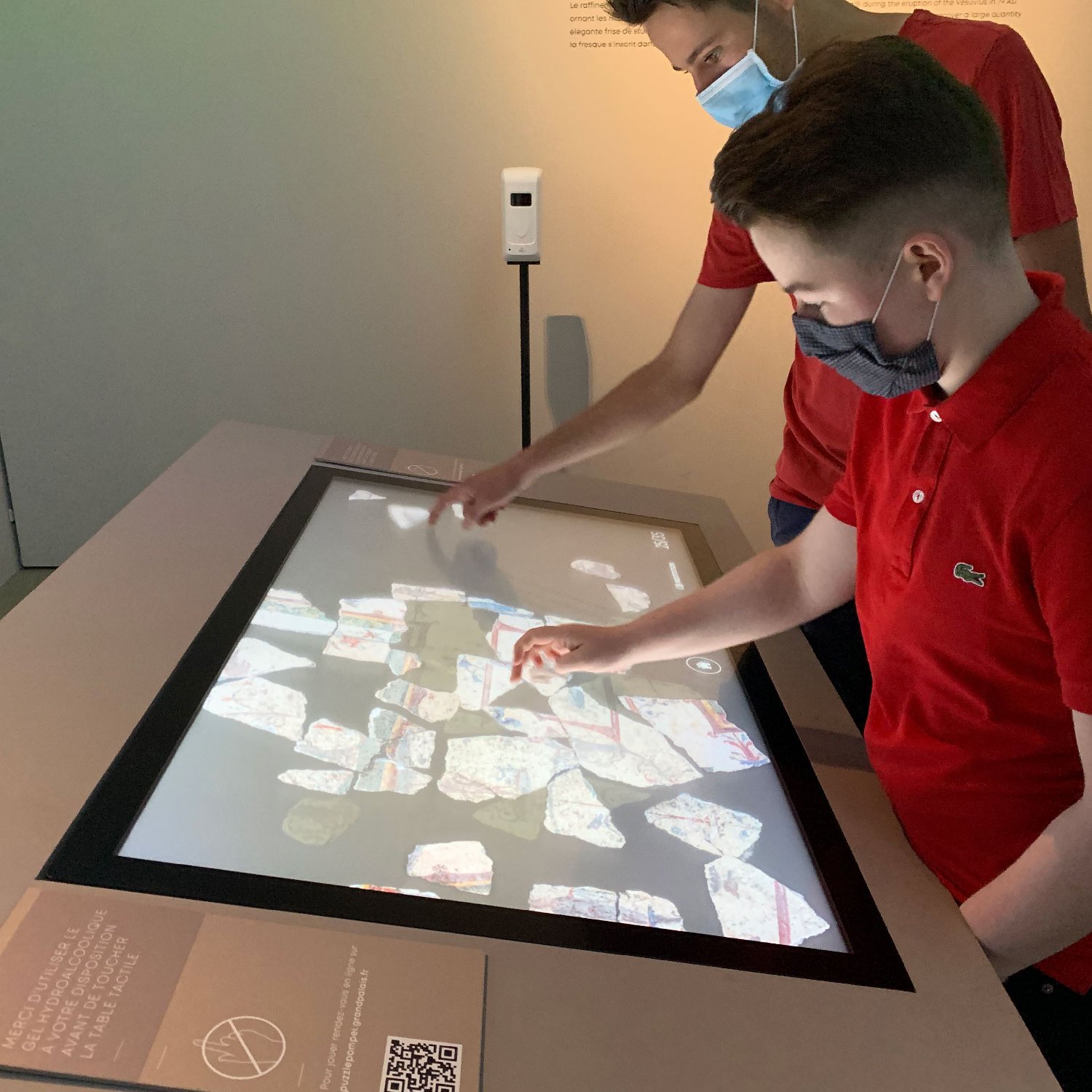 L'image montre deux jeunes garçons en train d'interagir avec un panneau tactile. Ils portent des masques et sont habillés en t-shirts rouges. Le panneau affiche des images de morceaux de pierres ou de fragments, probablement dans un contexte éducatif ou muséal, où ils explorent et découvrent des informations. L'ambiance semble moderne et engageante, favorisant l'apprentissage interactif.