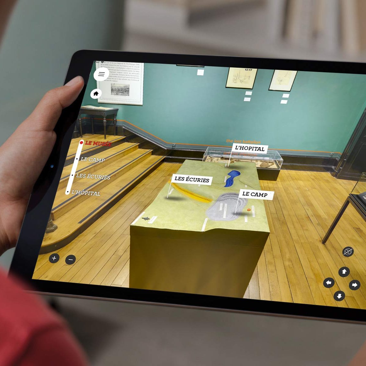 L'image montre une personne tenant une tablette, sur laquelle est affichée une application de réalité augmentée. Sur l'écran, on peut voir des éléments interactifs en rapport avec une exposition, avec des étiquettes comme "Les découvertes" et "Le camp". Le fond de l'image représente un musée, avec des murs verts et un sol en bois. La personne semble explorer des informations ou des objets exposés, mettant en avant l'utilisation de la technologie pour enrichir l'expérience muséale.