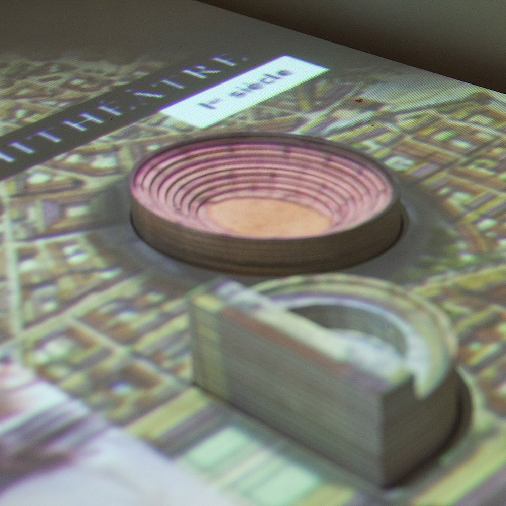 L'image montre une projection d'un amphithéâtre, probablement romain, avec des représentations en 3D. On peut voir des couches concentriques qui illustrent l'intérieur de l'amphithéâtre, ainsi qu'une vue d'ensemble de la zone environnante. La projection semble être sur une surface plane, donnant une idée claire de la structure et de son emplacement dans le contexte urbain.