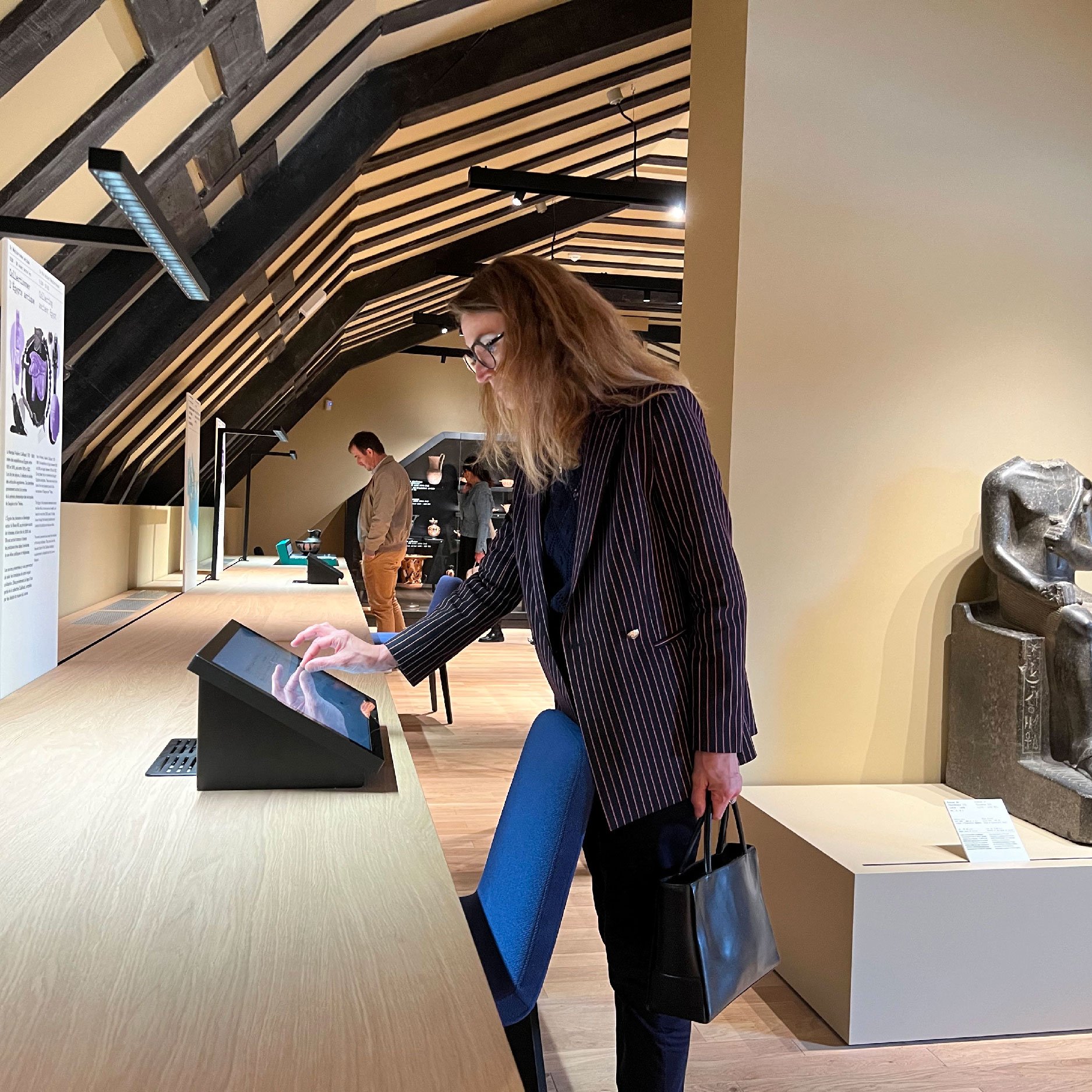 L'image montre une femme se tenant près d'un comptoir dans un musée. Elle semble interagir avec un écran tactile. En arrière-plan, on peut apercevoir d'autres visiteurs et une statue ou une sculpture ancienne sur un socle. La pièce a des poutres en bois au plafond, créant une ambiance chaleureuse et accueillante. La femme porte une tenue professionnelle avec un sac à main.