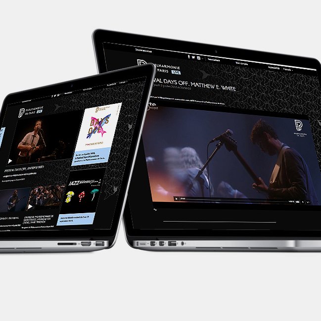 L'image montre deux ordinateurs portables inclinés, affichant des écrans avec ce qui semble être du contenu vidéo et des informations sur un artiste ou un événement musical. L'un des écrans montre un clip ou une performance, tandis que l'autre pourrait contenir des détails supplémentaires ou des panels. Le design des écrans est élégant et moderne, avec un fond noir et une interface stylisée.