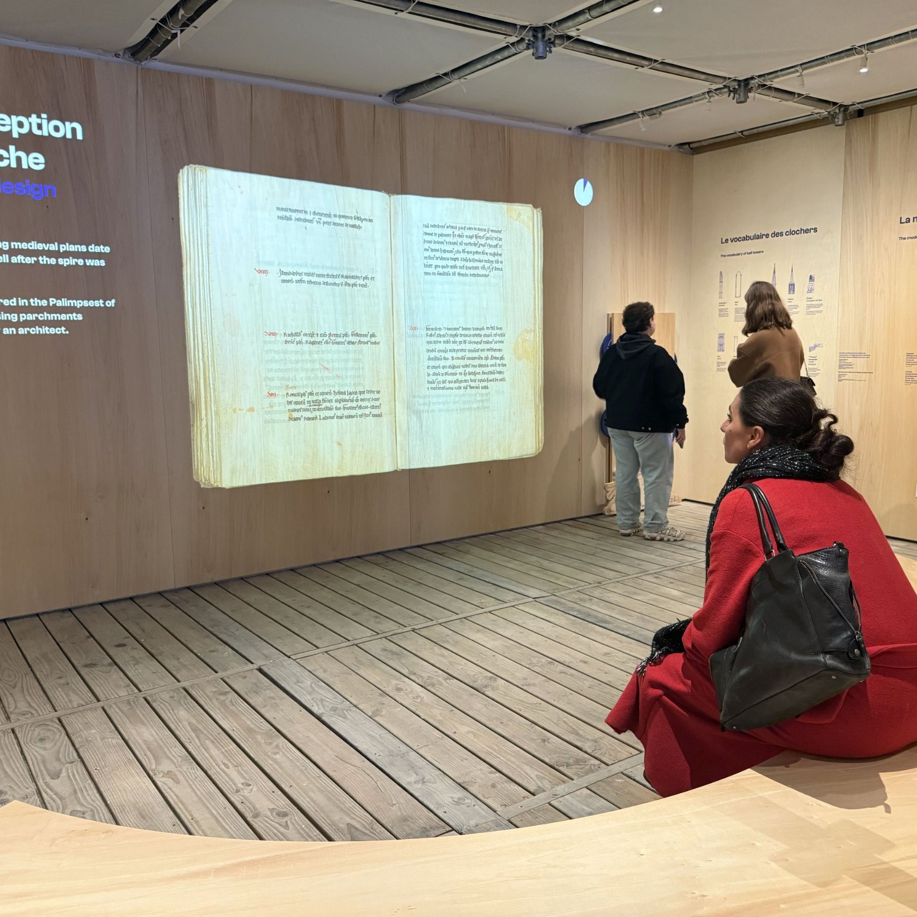 L'image montre une exposition dans un musée, où des visiteurs explorent des panneaux d'information. Au centre, on voit une femme assise sur une structure en bois, vêtue d'un manteau rouge et d'un sac à main noir. Les panneaux sur le mur incluent des informations sur le design des flèches architecturales, avec des illustrations et du texte en français. L'ambiance est moderne et épurée, avec des murs en bois et un sol en bois.