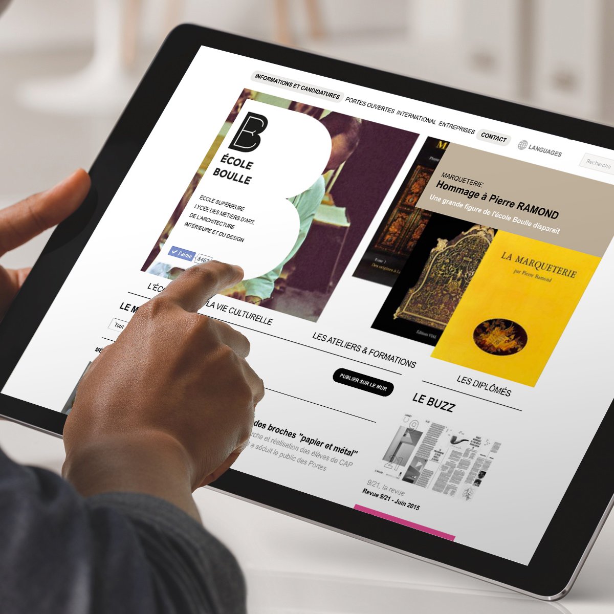 L'image montre une personne utilisant une tablette pour naviguer sur un site web. L'écran présente des informations sur une école, incluant des publications et des événements. On peut voir des visuels de livres et des sections comme "les expositions" et "le buzz". L'utilisateur semble intéressé par le contenu, interagissant avec l'écran tactile.