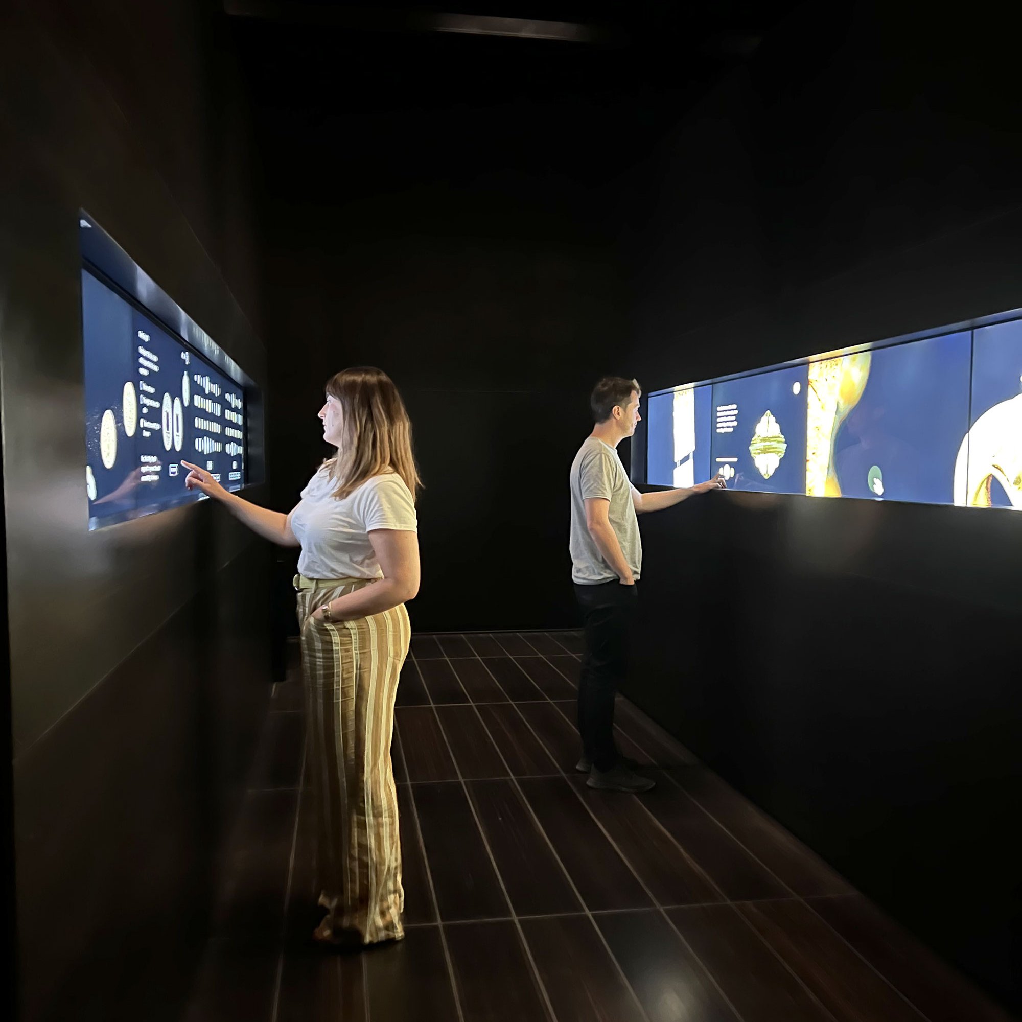 L'image montre deux personnes se tenant dans un espace intérieur sombre. Elles interagissent avec des écrans disposés sur les murs, qui affichent des informations visuelles. La femme, à gauche, porte un t-shirt blanc et un pantalon à rayures, tandis que l'homme, à droite, est vêtu d'un t-shirt gris et de jeans clairs. L'éclairage est subtil, créant une ambiance moderne et immersive.