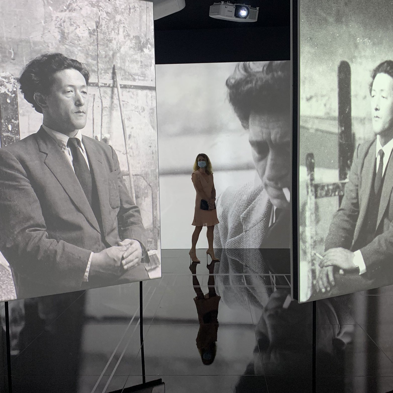 L'image présente une installation artistique moderne où une silhouette féminine se tient au centre d'un espace d'exposition. Cette personne porte un masque et se trouve entre plusieurs écrans qui affichent des photographies en noir et blanc. Ces images montrent un homme dans des poses réfléchies, évoquant une ambiance nostalgique et contemplative. La lumière des projections crée des reflets sur le sol, ajoutant une dimension dynamique à l'ensemble de la scène.