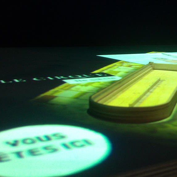 L'image montre une surface sur laquelle se trouve un livre ouvert, avec un accent sur le titre "Le Cirque". À côté, il y a un objet allongé, qui semble être un modèle ou une maquette, et une projection indique "VOUS ÊTES ICI !", suggérant une sorte d'interaction ou d'orientation dans un espace donné. L'ensemble est éclairé de manière à créer une atmosphère immersive et interactive.