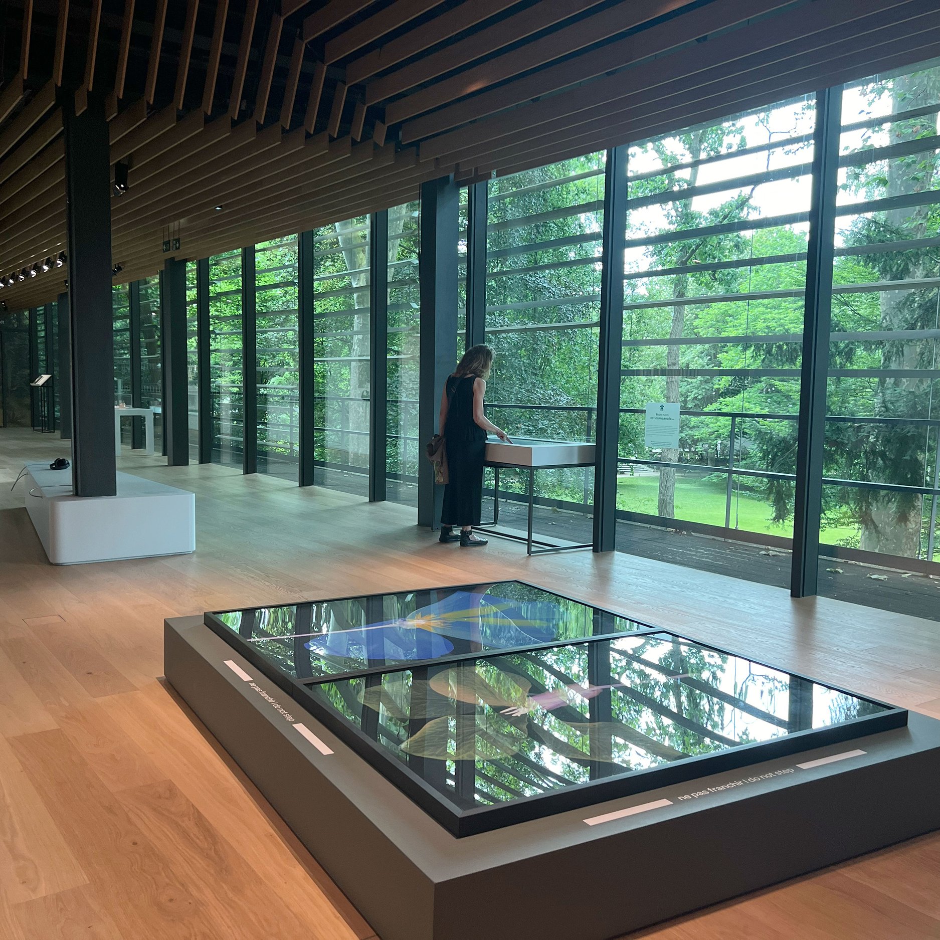L'image montre l'intérieur d'un musée ou d'une galerie d'art, avec un sol en bois clair et de grandes fenêtres qui laissent entrer la lumière naturelle. À gauche, on aperçoit une personne debout près d'une table, semblant observer une œuvre d'art ou une exposition. Au centre, il y a une grande installation ou un tableau en verre qui présente des couleurs vives. L'environnement est verdoyant, offrant une vue sur l'extérieur avec des arbres et de la végétation. L'ensemble donne une impression de modernité et d'harmonie entre l'art et la nature.