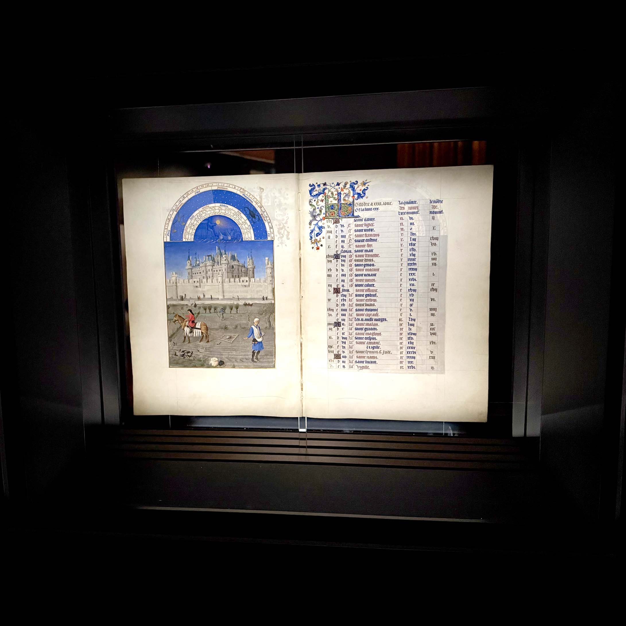 L'image montre un ancien manuscrit exposé dans une vitrine. Sur la gauche, une illustration colorée représente une scène avec des personnages, probablement à l'extérieur, devant un bâtiment imposant qui pourrait être un palais ou une cathédrale. Sur la droite, on trouve une liste écrite, probablement des noms ou des événements, ornée d'initiales décoratives en couleur. Le fond est sombre, mettant en valeur le manuscrit et ses détails complexes.