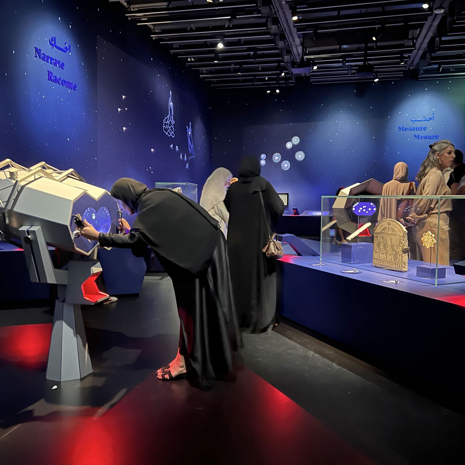 L'image montre une exposition dans un musée, probablement dédiée à l'astronomie ou à la science. Des visiteurs, dont plusieurs femmes portant des hijabs, interagissent avec des appareils de visualisation. En arrière-plan, on peut voir des objets exposés sous une lumière douce et une décoration étoilée sur les murs. L'ambiance semble à la fois éducative et attrayante, avec des présentations mettant en valeur des instruments scientifiques ou des artefacts culturels.