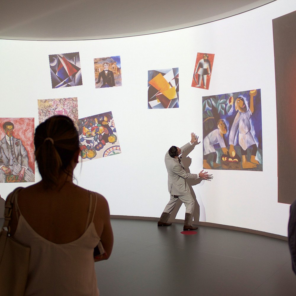 L'image montre une exposition artistique où un présentateur, habillé en costume clair, interagit avec des œuvres d'art projetées sur un mur courbe. Il semble expliquer ou mettre en valeur les installations, tandis qu'un groupe de visiteurs l'observe attentivement. Les œuvres affichées comprennent divers styles, tels que des peintures colorées et abstraites. L'ambiance est dynamique et immersive, illustrant une rencontre entre l'art et le public.