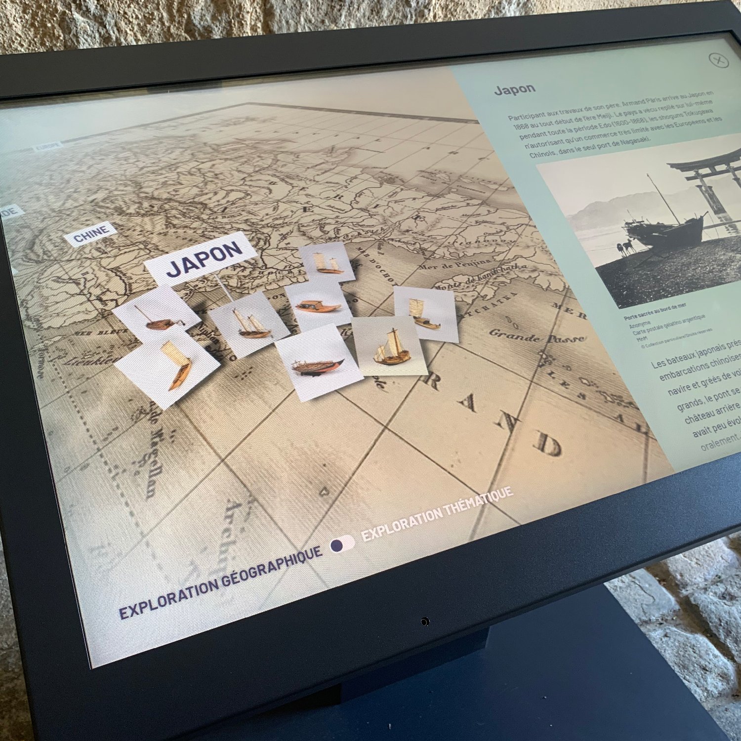 L'image montre un écran tactile interactif présentant une carte géographique du Japon, avec des éléments disposés sur cette carte. On peut voir des informations textuelles sur le Japon sur le côté droit de l'écran, ainsi que des icônes représentant des objets ou thèmes de l'exploration. Le fond de l'écran a un motif qui ressemble à une carte ancienne. Le tout semble être installé dans un environnement muséal avec un sol en pierre.