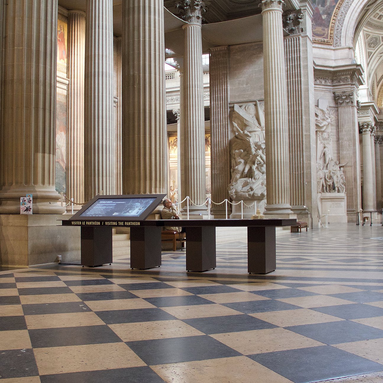 L'image montre l'intérieur d'un grand bâtiment aux colonnes majestueuses, probablement un monument historique comme un panthéon ou une cathédrale. Le sol est composé de carreaux en damier, alternant entre des teintes claires et sombres. Au centre, il y a une table interactive avec un écran. Des sculptures et des œuvres d'art sont visibles sur les murs, et l'éclairage met en valeur l'architecture impressionnante de l'endroit. Des visiteurs se déplacent tranquillement dans l'espace.