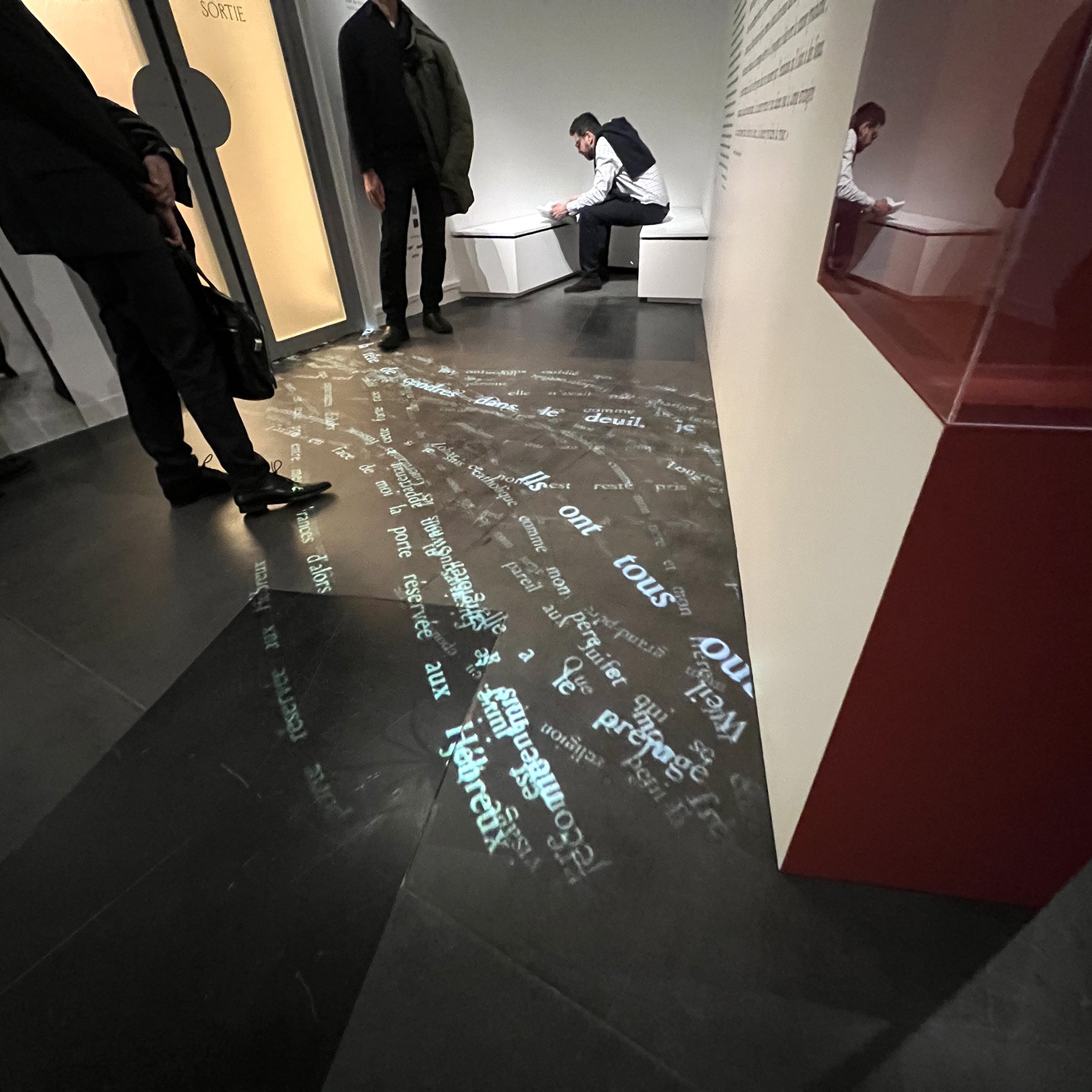 L'image montre un espace intérieur, probablement un musée ou une galerie. Au sol, des mots et des phrases sont projetés, créant un motif lumineux sur le sol. On peut voir des silhouettes de personnes, certaines debout, d'autres assises, interagissant avec l'environnement. Les murs semblent comporter des inscriptions ou des informations, mais elles ne sont pas clairement visibles dans l'image. L'atmosphère semble être moderne et intellectuelle, réfléchissant une intention artistique ou informative.