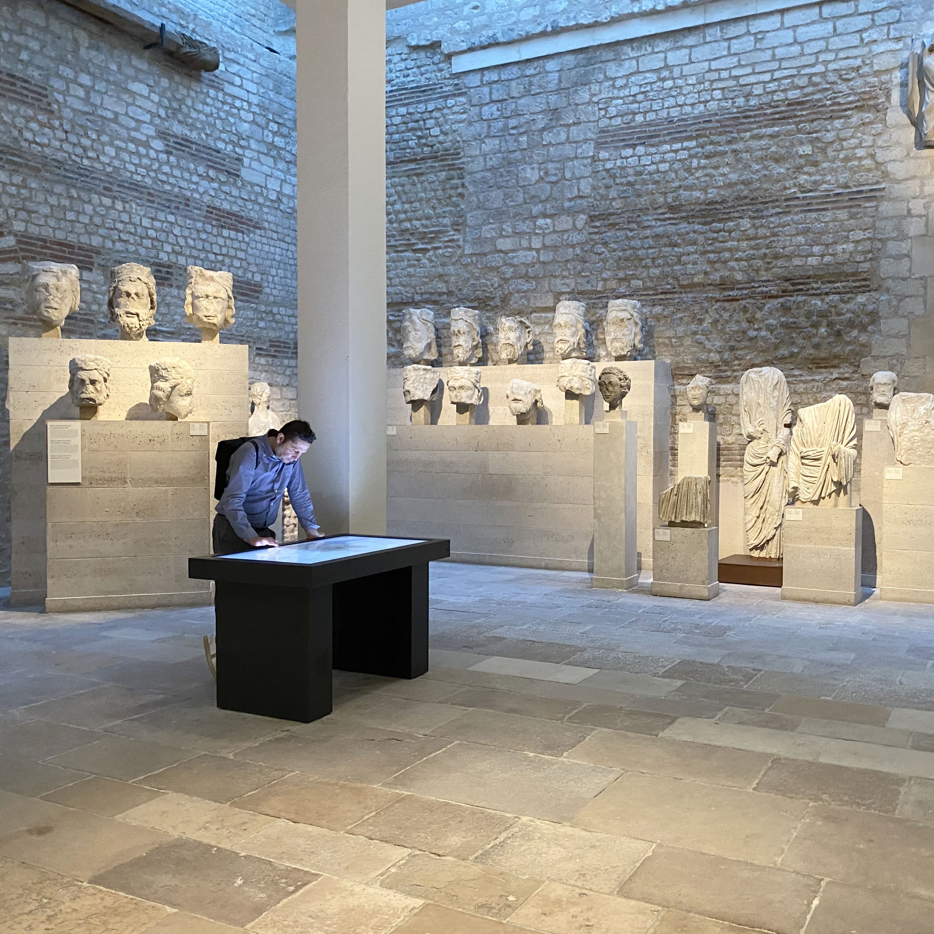L'image montre une salle d'exposition avec des sculptures anciennes. À gauche, plusieurs bustes en pierre sont disposés sur des socles, tandis qu'à droite, une autre sculpture intrigante est visible. Un homme est penché sur une table noire au centre de la pièce, probablement en train d'examiner un document ou une carte. La lumière naturelle entre à travers des fenêtres, créant une atmosphère calme et contemplative, accentuée par les murs en pierre apparente.
