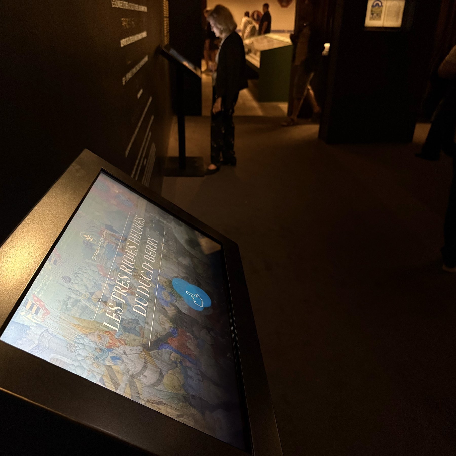 L'image montre un dispositif interactif, probablement un écran tactile, monté sur un support. L'écran affiche une interface numérique, peut-être dédiée à une exposition ou à une présentation. On peut voir du texte et potentiellement des images ou des graphiques sur l'écran. Le fond est sombre, ce qui met en valeur l'écran. À proximité, il y a un panneau explicatif sur le mur, qui pourrait fournir des informations supplémentaires sur le sujet présenté.