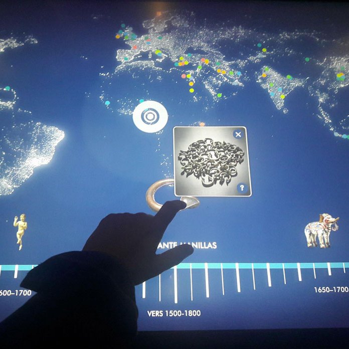 L'image montre un écran interactif où une main touche un point sur une ligne du temps. Cette ligne du temps s'étend de 1500 à 1800, avec différentes étapes, représentées par des illustrations variées, qui semblent indiquer des événements ou des thèmes historiques. En arrière-plan, on voit une carte du monde avec des points colorés, probablement pour indiquer des localisations spécifiques. Les éléments affichés peuvent inclure des objets ou des symboles liés à des cultures, des civilisations ou des découvertes.
