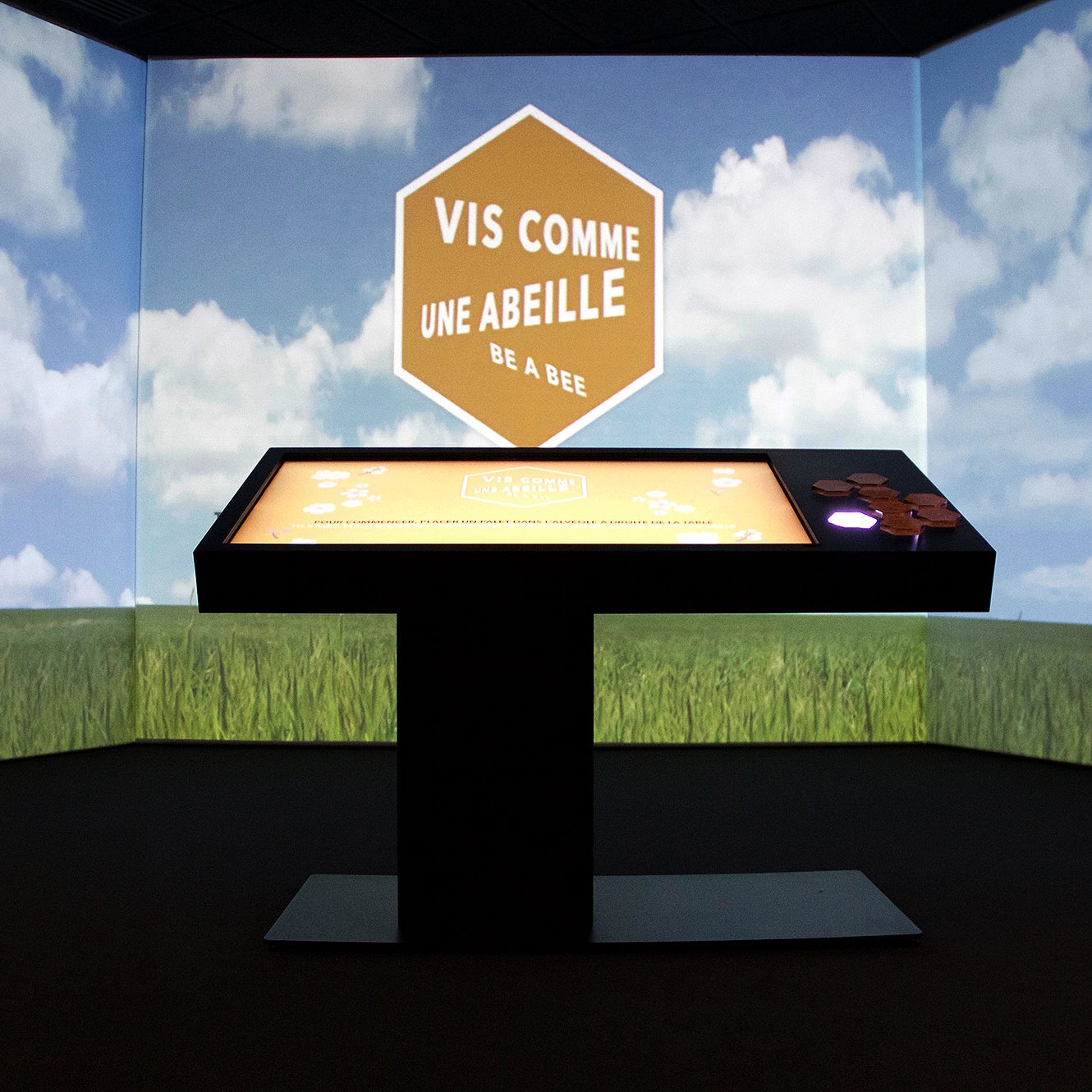 L'image montre une installation interactive dans une salle, avec un écran de projection qui affiche un ciel bleu avec des nuages. Au centre, il y a une table tactile noire sur laquelle se trouve le texte "VIS COMME UNE ABEILLE" en orange, accompagné de la phrase "be a bee". L'environnement dégage une atmosphère ludique et éducative, suggérant un thème lié aux abeilles et à leur comportement. Les murs projetent des images de nature, comme des champs de verdure.