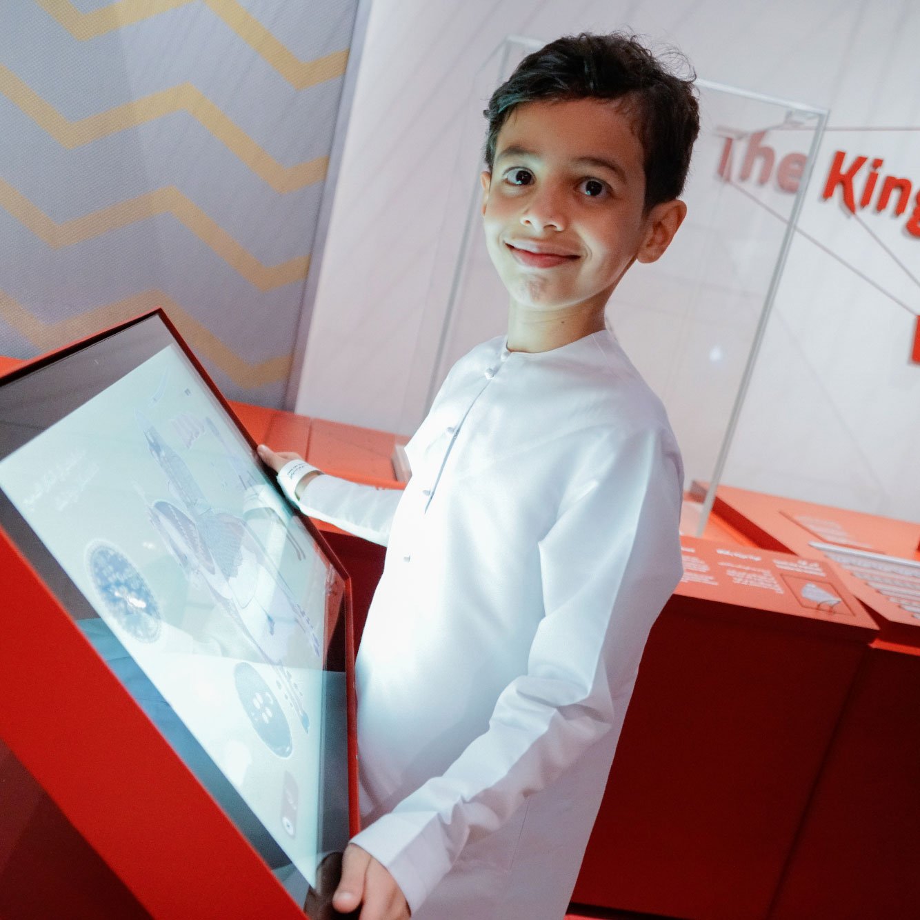 L'image montre un jeune garçon souriant, habillé d'une tenue traditionnelle blanche. Il se tient devant un écran interactif dans un environnement exposé, probablement dans un musée. L'interface de l'écran semble afficher des informations ou des graphiques. L'arrière-plan est minimaliste, avec des formes géométriques et des couleurs vives, principalement du rouge.