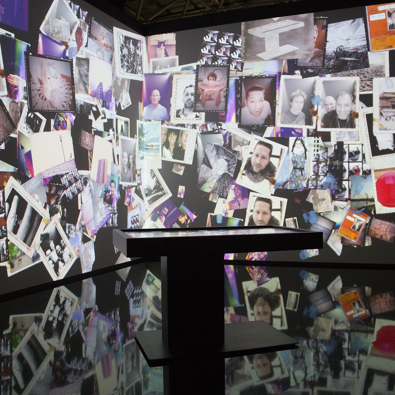 L'image montre une installation artistique immersive où des photographies sont projetées sur les murs. Ces images sont disposées de manière disparate, créant une mosaïque visuelle. On peut voir une variété de photos, dont certaines semblent être des portraits et d'autres des paysages ou des objets. Le sol est noir et réfléchissant, ajoutant une dimension supplémentaire à l'ensemble. L'atmosphère un peu sombre et l'accumulation de ces souvenirs visuels semblent inviter à une réflexion personnelle ou à une exploration du passé.