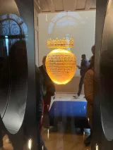 L'image montre un objet suspendu dans une vitrine, probablement un bijou ou un artefact. L'objet a une forme ronde et est décoré de motifs raffinés, avec un effet lumineux qui évoque un matériau précieux, comme de l'or ou du verre poli. Une couronne délicate est visible sur le dessus de l'objet, ajoutant à son allure majestueuse. En arrière-plan, on aperçoit des silhouettes de visiteurs, suggérant que cet objet est exposé dans un musée ou une galerie.