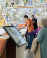L'image montre une scène dans un espace d'exposition ou un musée. Au premier plan, une femme d'âge mûr est debout, observant quelque chose, tandis qu'au centre, deux hommes interagissent avec des écrans ou des tableaux informatifs. L'homme à gauche porte un t-shirt orange et semble concentré, tandis que l'autre homme, en arrière-plan, porte une chemise sombre. L'arrière-plan est décoré de grandes illustrations colorées, suggérant un environnement artistique ou éducatif.