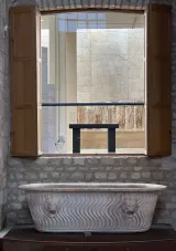 L'image montre une salle d'exposition avec un grand bain en marbre, orné de motifs en forme de vagues et de têtes de lions aux extrémités. Ce bain est placé sur une plateforme en bois sombre, entouré de murs en pierre claire. À l'arrière-plan, on aperçoit une vaste fenêtre ouverte qui laisse entrer la lumière naturelle, éclairant le bain. À travers cette fenêtre, on peut voir des éléments d'autres expositions, suggérant un environnement muséal. Les volets en bois de la fenêtre sont ouverts, ajoutant une touche classique à la scène.