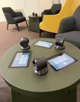L'image montre une table ronde de couleur verte avec quatre écrans tactiles intégrés. Chaque écran est entouré de dispositifs ronds noirs, probablement des stations d'écoute ou des contrôles. En arrière-plan, on peut apercevoir des fauteuils confortables en divers coloris, comme un jaune et un gris. L'ensemble semble être dans un espace moderne et accueillant, suggérant une ambiance de détente ou de consultation.