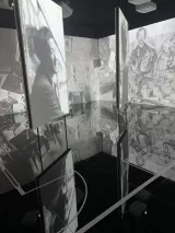 L'image montre une installation artistique immersive dans un espace sombre. Des panneaux surdimensionnés sont disposés autour de la pièce, affichant des photographies et des illustrations en noir et blanc. Le sol en miroir reflète ces images, créant un effet visuel intrigant et dynamique. L'ensemble de l'installation semble axé sur la relation entre l'art et la mémoire, avec des éléments de collage ou de esquisse en arrière-plan.