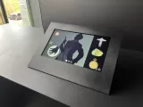 L'image montre un écran interactif monté sur une surface noire. L'écran présente une silhouette d'un homme, habillé d'un costume élégant, tenant un objet dans une main. À côté de la silhouette, plusieurs illustrations d'objets sont visibles, dont une montre de poche et d'autres éléments qui pourraient être des artefacts ou des bijoux. L'environnement semble moderne, avec des fenêtres laissant entrer la lumière naturelle.