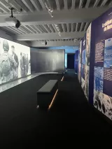 L'image montre une exposition intérieure, probablement dans un musée. On peut voir un espace bien éclairé, avec des murs décorés de panneaux informatifs, mettant en avant des événements ou des thèmes historiques. Sur le mur à gauche, des photographies en noir et blanc semblent illustrer des scènes historiques, tandis que le sol est recouvert d'un revêtement sombre. Au centre de l'espace, un banc gris est présent, invitant à la contemplation. L'atmosphère générale est moderne et sobre, invitant à la réflexion sur le sujet exposé.