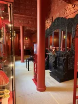 L'image montre une pièce intérieure ornée de motifs détaillés. Les murs et le plafond sont de couleur rouge vif, avec des décorations sculptées. On y voit des colonnes également rouges. À droite, il y a un mobilier noir, comprenant de grandes chaises ou un trône, et à gauche, des vitrines présentant divers objets, probablement des artefacts culturels. Le sol est recouvert d'un matériau clair, ajoutant un contraste avec les couleurs riches des murs et du mobilier. L'ensemble de la pièce dégage une atmosphère traditionnelle et raffinée.