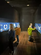 L'image montre une salle d'exposition ou un musée, avec des murs sombres et des écrans interactifs au niveau des yeux. Quatre personnes sont visibles, certaines en train d'interagir avec les écrans. L'une des personnes porte un manteau orange, une autre a les cheveux longs et blonds, et il y a aussi des individus habillés de manière décontractée. L'éclairage de la pièce est subtil, créant une atmosphère moderne et immersive.