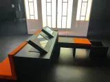 L'image montre un espace intérieur avec des meubles modernes, notamment des tables et des sièges. Les tables sont rectangulaires et possèdent des écrans intégrés, tandis que les bancs sont recouverts d'un tissu orange et noir. L'environnement semble sombre, ce qui donne un aspect contemporain et minimaliste, et il y a un éclairage en arrière-plan qui rappelle des portes ou des fenêtres. Les couleurs dominantes sont le noir et l'orange, créant un contraste visuel marqué.