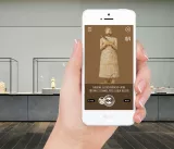 L'image montre une main tenant un smartphone affichant une application. L'écran de l'application invite l'utilisateur à chercher des différences entre une sculpture représentée et une image fournie. En arrière-plan, on aperçoit une exposition dans un musée, avec plusieurs vitrines contenant des objets d'art ou des bijoux, et un environnement sobre et moderne.